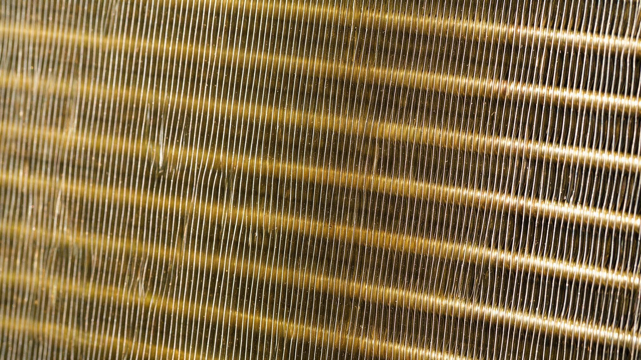 A rectangular block of closely spaced metallic gold aluminium fins with copper U-bends and open copper tube ends, beaded with water droplets, stands on a glossy navy studio floor with a soft reflection while two thin translucent pale-blue air ribbons curve across it.