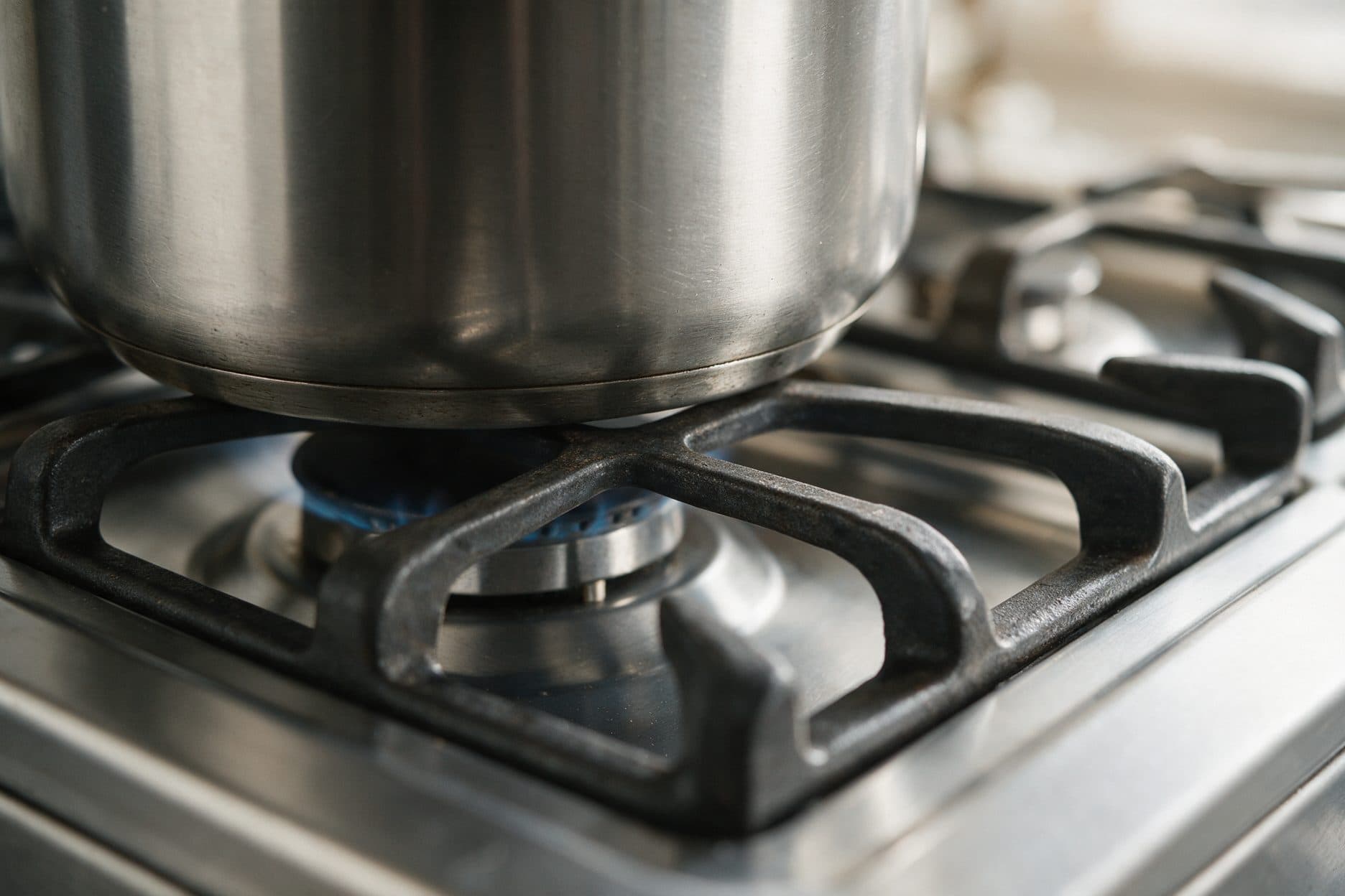 A stainless steel pot sits on a cast iron burner grate over a lit gas flame, shown close up on a stove.