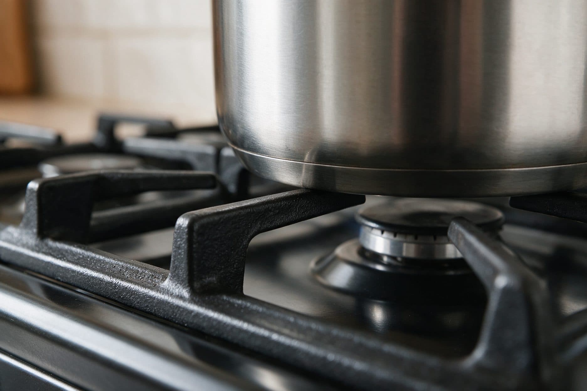 A dark stainless stockpot sits on a cast iron burner grate, its bottom edge resting directly on the trivets arms with a warm sunlit kitchen blurred in the background.