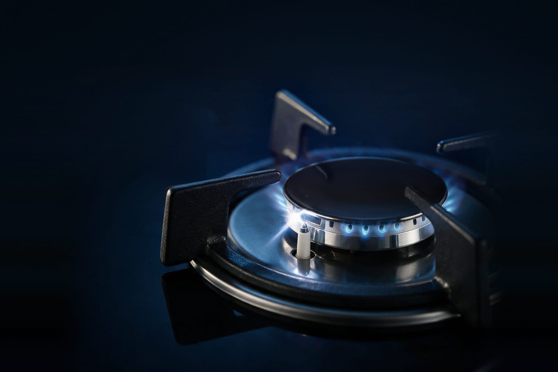 A gas burner on a black glass hob is lit with a ring of small blue flames under a flat black cap, a white spark flickers at the ignition electrode, and dark cast-iron pan supports surround it against a deep navy background.