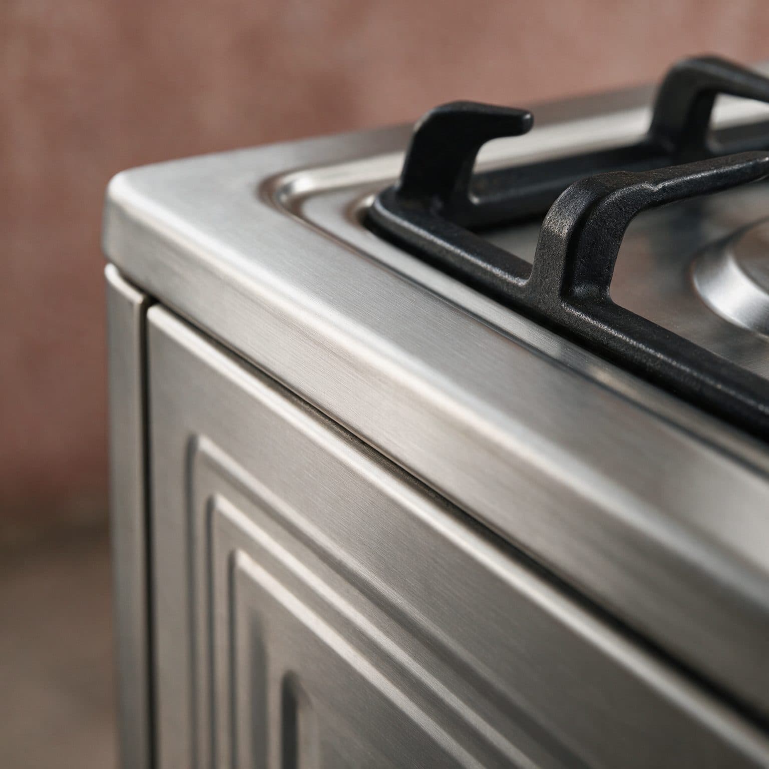 A close-up macro shows the corner of a stainless-steel gas-cooker worktop with two cast-iron burner grates, a black control knob and the top edge of the closed oven door, lit by soft warm window light in a home kitchen.