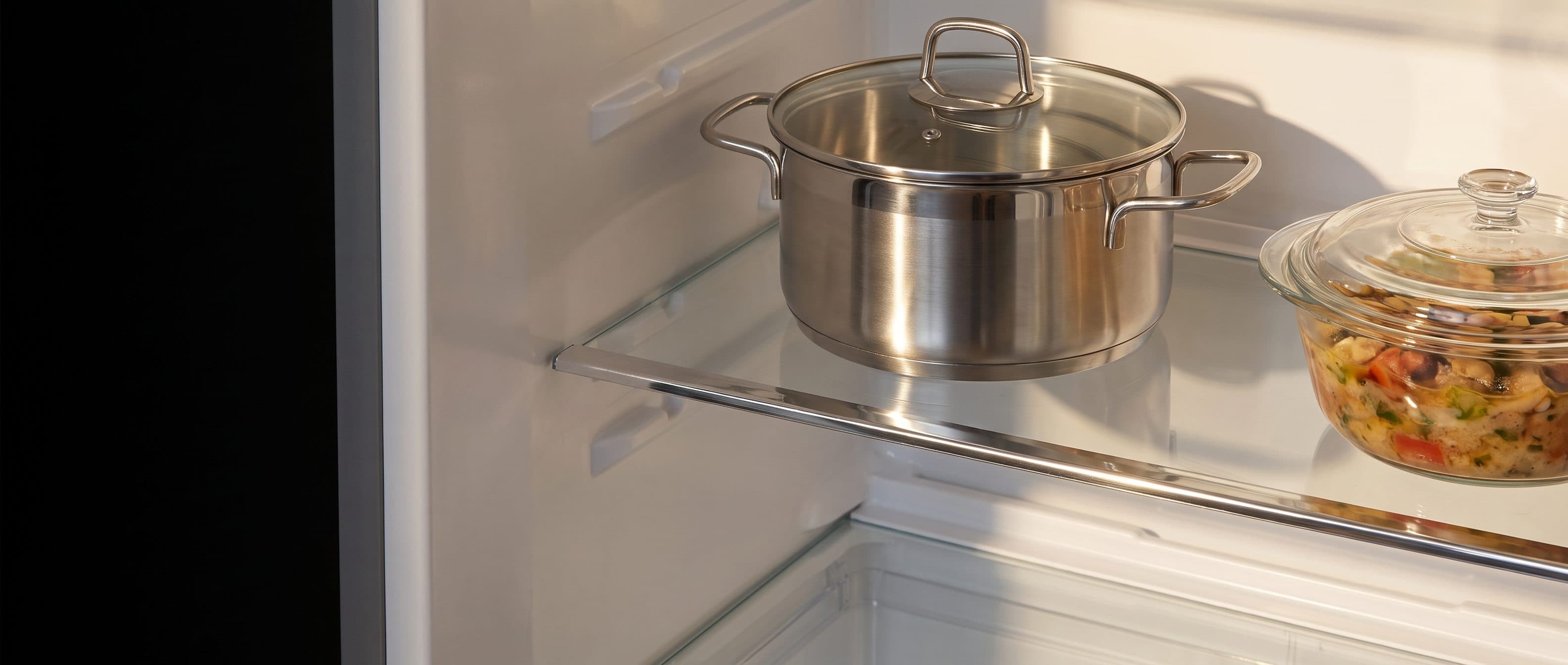 A large lidded stainless steel pot standing on a clear glass refrigerator shelf, the polished edge of the glass lit along its length.