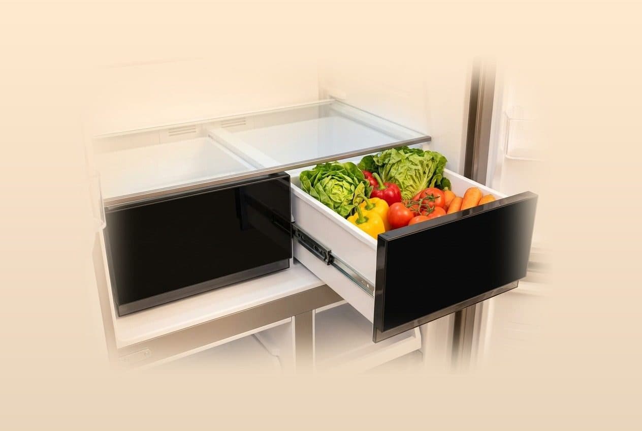 Two black-fronted fridge crisper drawers sit side by side in a warm cream-toned interior, the right one slid out on metal runners and filled with lettuce, red and yellow peppers, tomatoes and carrots.