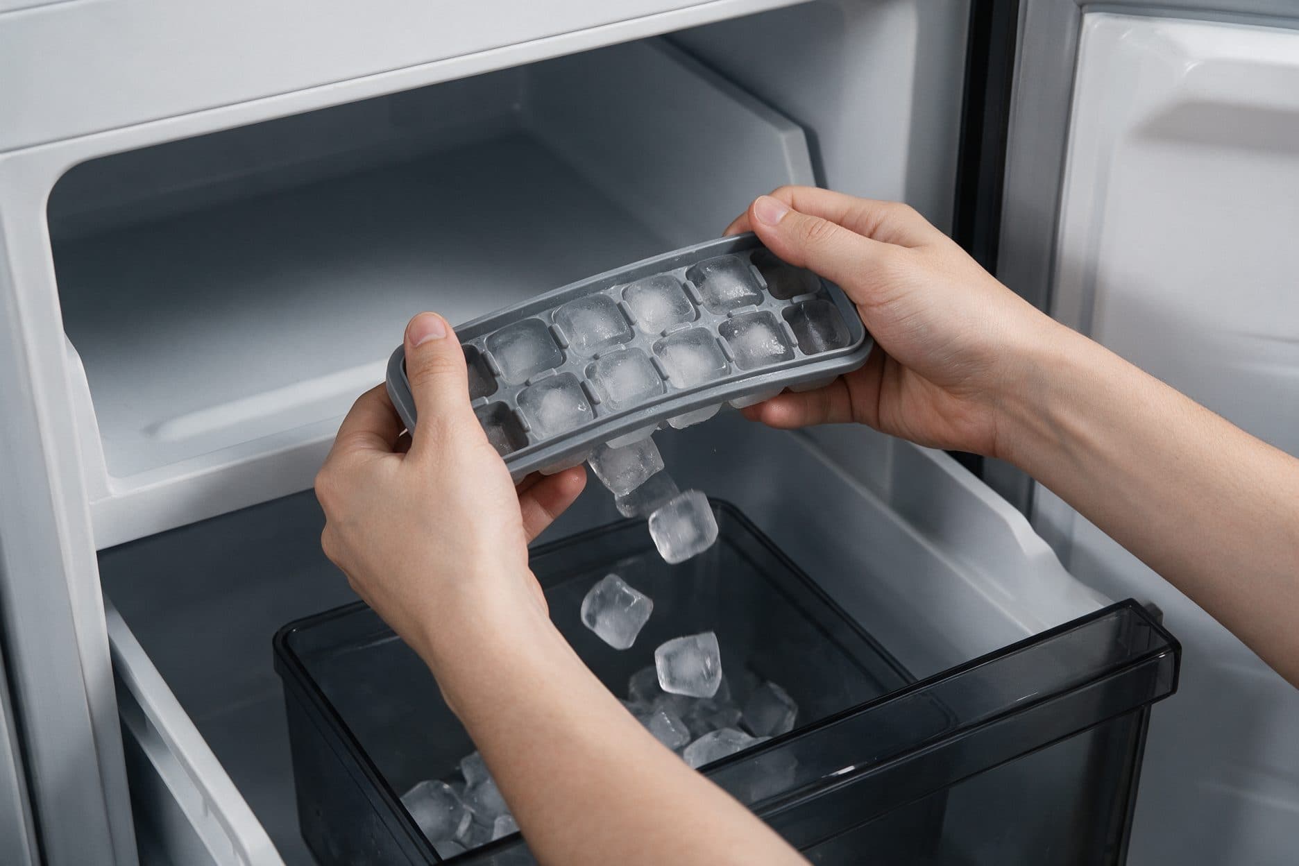 Two hands twist a flexible ice tray over an open freezer ice bin, with cubes dropping out of the tray into the black bin below, shot inside a white-and-grey freezer compartment.