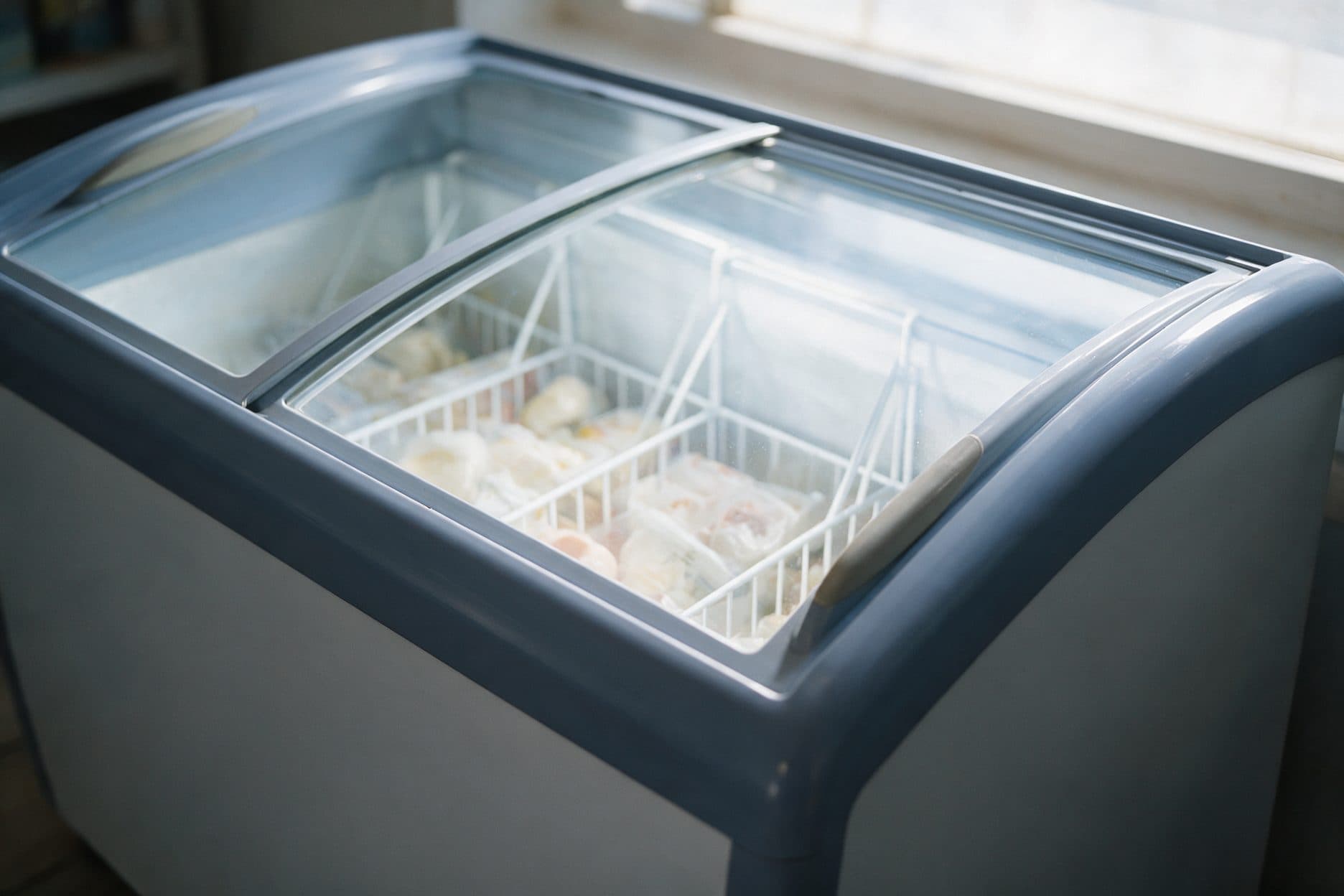 A chest freezer with two sliding curved-glass lids sits in a sunlit room, one lid open to reveal a wire storage basket with dividers holding plain-wrapped frozen food, soft window light falling across the frosted glass.