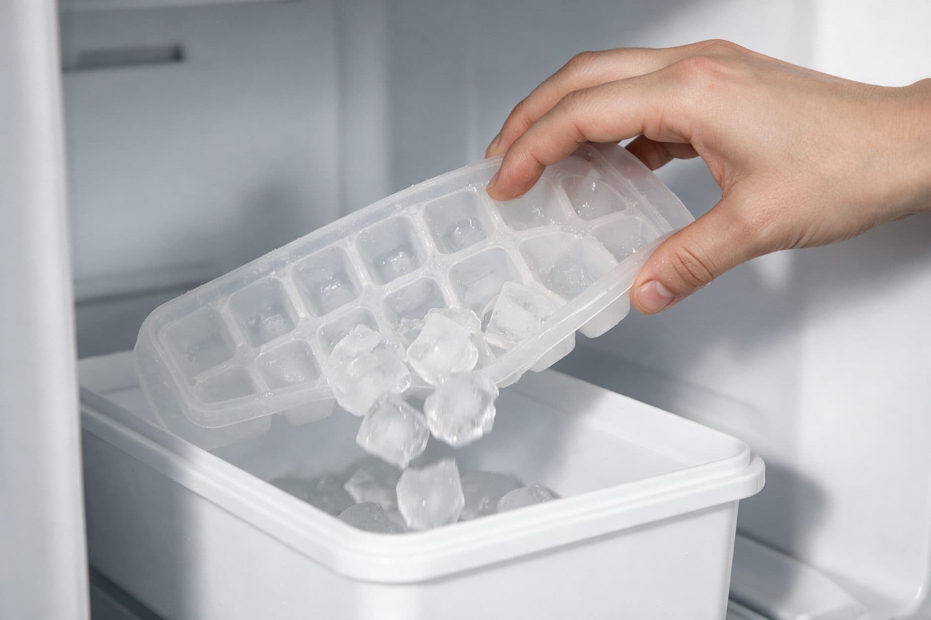 A hand tilts and twists a plastic ice cube tray over a white storage box inside an open freezer compartment, with ice cubes dropping from the tray into the box.